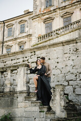 Fototapeta na wymiar Woman in black long dress hugs man stand on stairs near ancient palace. Female kissing male in street. Luxury couple newlyweds near old Pidhirtsi Castle, Lviv, Ukraine. Stylish bride and groom outdoor