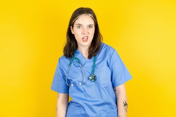 Portrait of dissatisfied Young caucasian doctor woman wearing blue medical uniform smirks face, purses lips and looks with annoyance at camera, discontent hearing something unpleasant - obrazy, fototapety, plakaty