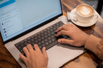 A guy is typing in a chat on laptop and coffee cup and saucer kept aside on wooden table