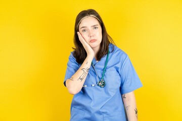 Sad lonely Young caucasian doctor woman wearing blue medical uniform touches cheek with hand bites...