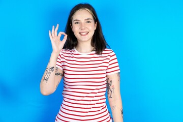 beautiful young woman wearing striped T-shirt hold hand arm okey symbol toothy approve advising...
