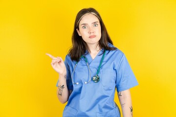 Serious Young caucasian doctor woman wearing blue medical uniform smirks face points away on copy space shows something unpleasant. Look at this advertisement. Big price concept