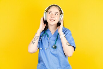 Young caucasian doctor woman wearing blue medical uniform wears stereo headphones listens music concentrated aside. People hobby lifestyle concept