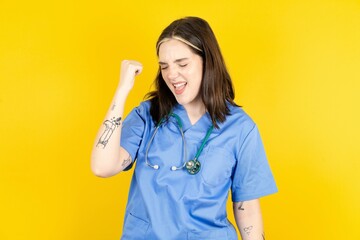 Portrait of funny Young caucasian doctor woman wearing blue medical uniform shout yeah raise fists...