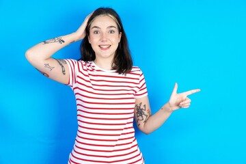 Surprised beautiful young woman wearing striped T-shirt pointing at empty space holding hand on head