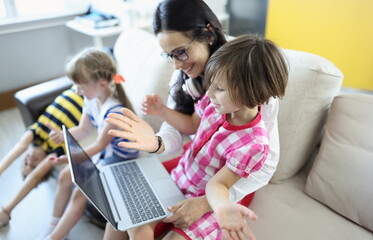 Woman is sitting on the couch, girl is sitting on her lap, they are looking at laptop next to children are playing. Education quarantined children online concept