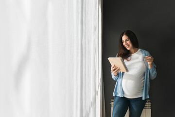 Smiling pregnant woman standing with notebook at home