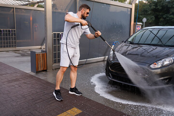 A man washes the front grill of a car with a water cannon. A car at a self service car wash.