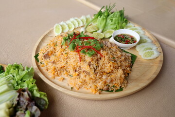 Pork fried rice with egg topped with coriander leaves and sliced ​​red chili. Fried rice served on a wooden plate decorated with sliced ​​cucumbers, lime wedges, spring onions and chili fish sauce.

