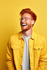 Obraz premium Portrait of a laughing handsome young man, with red hair, wearing spectacles and a yellow jacket, on a yellow background 