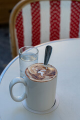 A cup of cappuccino and a glass of water on a white surface against the background of a red and white chair