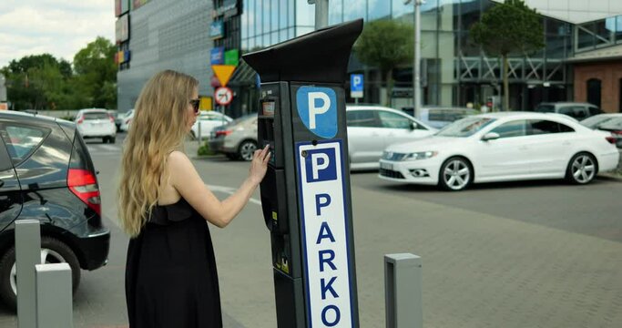 Young woman in sunglasses and summer dress approach parking meter and enter code. Beautiful female car driver with long hair come to outdoors parking machine and input plate number on warm sunny day.