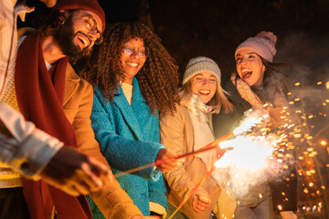 Young group of friends having fun together at night outdoors winter party holding bengal light with sparklers fireworks celebrating at the new year party 