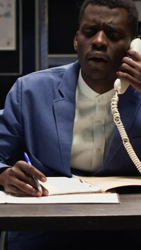 Vertical video African American detective and Caucasian criminologist gather evidence and examine files. Male investigator answering landline phone, receiving important clues and information to solve