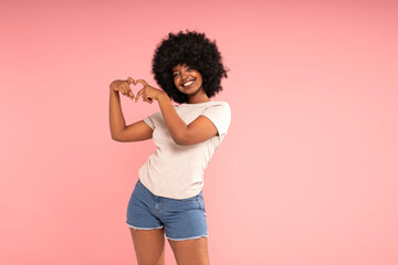 Optimistic african american woman showing heart sign to the camera, smiling happily.  Girl with afro hairstyle wearing casual tshirt and short jeans - 644894144