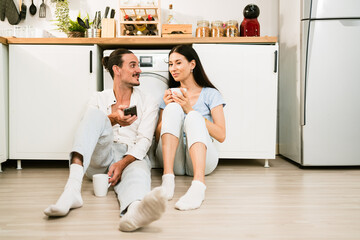 Happy Caucasian couple sitting together on the kitchen floor together while a man using a smartphone for social media or e-commerce with discussion. Young man and woman talking while at home.