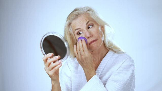 Mature middle aged woman 50s, 60s, applying natural makeup powder foundation with cosmetic sponge on clean face skin smoothing wrinkles, looking in mirror beauty routine white background.