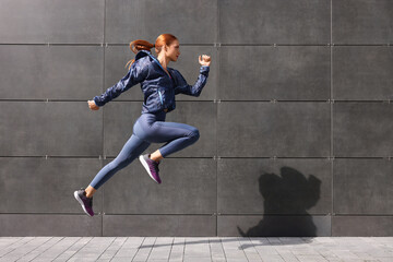 Beautiful woman in gym clothes jumping near dark grey wall on street, space for text