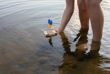 child, standing in a river, launches a home-made boat made of wine corks, instead of sail, a yellow-blue heart in colors of Ukrainian flag. Symbol of freedom, independence. Patriotic children's games
