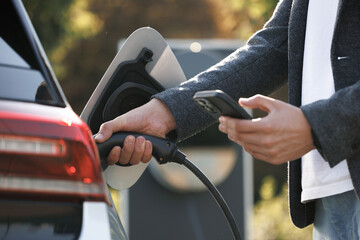Businessman charging electric car at outdoor charging station. Unrecognizable man plugging electric...