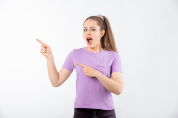 Surprised young woman with long hair points fingers at banner, shows great promo offer aside, looking amazed by promotion offer, stands in casual t shirt over white background