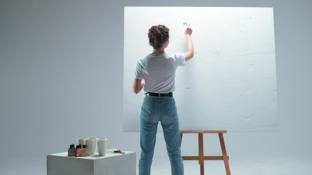 Back view, young woman artist covers a layer of white primer on a large canvas in a white room, a talented artist preparing to draw, abstraction.