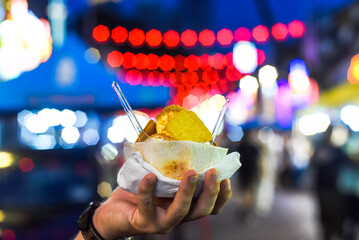 Obraz premium Hand holding coconut and mango ice cream in coconut shell against lights of Jalan Alor street food market in Kuala Lumpur