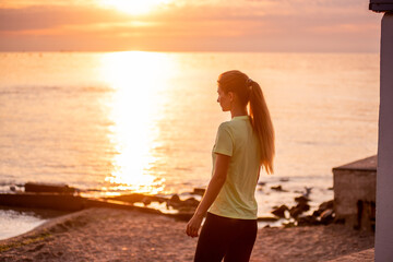 Portrait of young woman in sportswear who stopped for break between workouts. Sportswoman looks at dawn of sun on the sea. Girl walks along the embankment after her morning routine. Outdoor workout