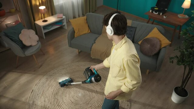 Slow motion video, from above joyful, happy young man vacuuming the carpet, wearing headphones and listening to music, dancing, then looking at the camera and smiling.