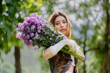 a young woman in a white dress stands on a green field and holds a large bouquet of flowers in her hands. In the background, there are forest and a lake. Lifestyle concept.