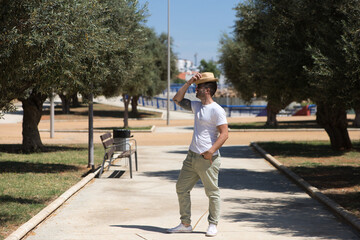 Handsome young man dressed in casual clothes with sunglasses and hat walks among the olive trees in a typical mediterranean style park in seville. The man is on holiday in Spain. Travel and tourism.