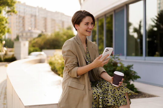 Outdoor photo of stylish beautiful woman with short haircut  enjoing autu,n day  holding cup of coffee, using mobyle phone. Weaering casual  jacker and elegant dress.
