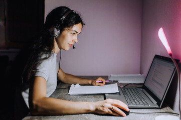 A young girl, a woman works at home, sitting on a laptop in headphones with a microphone at night, sitting at a table. Photography, portrait, lifestyle.