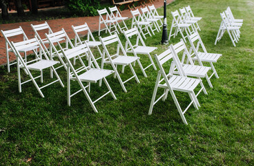 Lots of white wooden folding chairs line up on the green grass at an outdoor wedding ceremony.