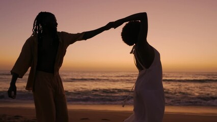 A black woman and African male, silhouetted against a mesmerizing sunset, walk on the beach, sharing smiles and flirtatious moments, radiating love and romance