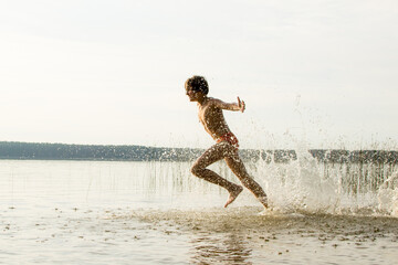 A slender guy child teenager runs along the water along the coast at sunset, splashing from the water.