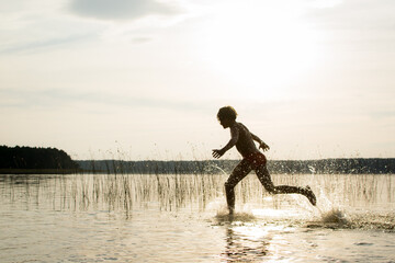 A slender guy child teenager runs along the water along the coast at sunset, splashing from the water.