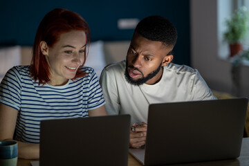 Man and woman work at computers together. Joint online remote work on Internet. Young African-American guy and Caucasian girl play along. Colleagues discuss project sitting at laptops at home. 