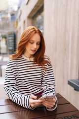Vertical portrait of pretty redhead young woman sitting outdoors cafe using smartphone chatting with friends looking to device screen. Beautiful female texting message on mobile phone in summer time.