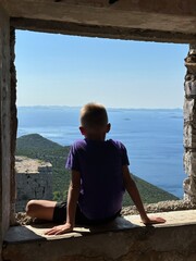 A child sits on the window of an old fortress. A teenager watches from the ruins of a fortress. The boy looks out the window at the seascape.
