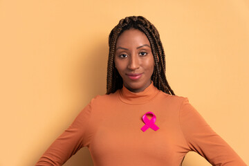 confident afro brazilian woman showing pink ribbon in beige background. breast cancer, awareness...