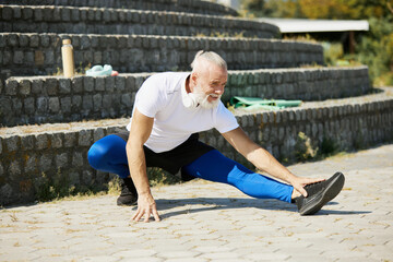 Sportive mature man in sportswear training outdoors on sunny day, doing legs stretching exercises. Concept of active and sportive lifestyle, age, health care, exercising, vitality, ad