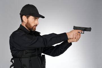 Focused bearded police officer taking aim, pointing with handgun in studio. Side view of caucasian cop shooting, holding gun, looking away, isolated on gray background. Concept of danger work, weapon.