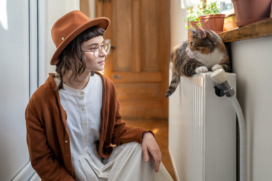 Interested woman cat owner in hat looks to fluffy pet lying on radiator under sunny window. Interaction domestic animal and mistress. Good attitude to environment joy of home. Inspiration from caring.