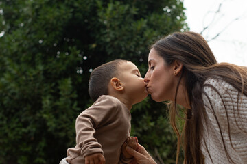 Fototapeta premium Mom kissing 1 year old baby on the mouth and holding his hand, a very tender image of true love. Autumn colors