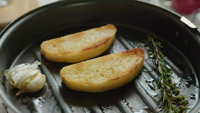 Hand of male chef turning baguette slices on grill pan while frying bread on olive oil with rosemary and garlic. Close-up impersonal shot