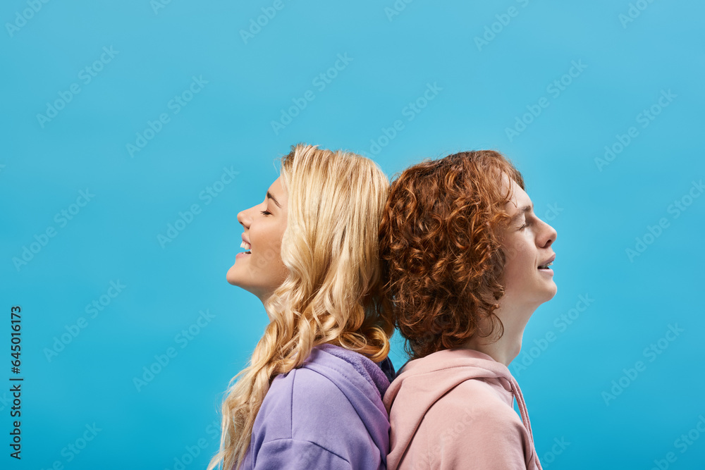 Wall mural carefree teenage friends sitting back to back and smiling with closed eyes on blue, side view