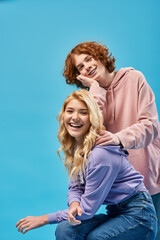 happy redhead teenage boy leaning on blonde girlfriend looking at camera and laughing on blue