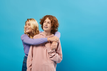 carefree teenagers in hoodies looking away on blue, happy blonde girl embracing teen guy, friendship