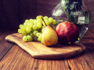 Still life image with green grapes, pear and pomegranate and glass bowl on wooden board and table, dark background. Selective focus.